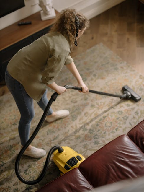 A middle-aged man with gray hair and glasses is vacuuming a yellow and orange patterned area rug in a bright living room with a modern aesthetic. The room features large windows with white trim and sh
