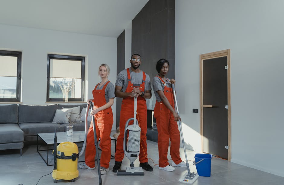A person dressed in a grey sweater and black pants is performing surface cleaning using a mop in a modern living room. The room features a grey sofa with cushions, a black side table, and large window