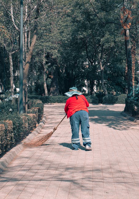 Two workers in navy uniforms with reflective stripes are engaged in surface cleaning and tidying up a paved pathway in a park area, using traditional straw brooms. They are surrounded by large trees w