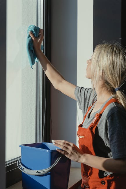 A woman with blonde hair tied back, wearing an orange apron and casual clothing, is cleaning a large glass window in a bright room using a blue cloth. She is standing indoors, holding a small blue bucket with a white handle in her left hand, while cleaning the window with her right hand. The window has a black frame, and the sunlight illuminates the scene, highlighting the clear, dust-free glass surface. The interior features neutral tones, with smooth walls and a clean, organized environment, reflecting the process of surface cleaning and maintenance as part of domestic cleaning services offered by Hounslow Cleaner. The image emphasizes thorough window cleaning and the importance of hygiene in a residential setting.