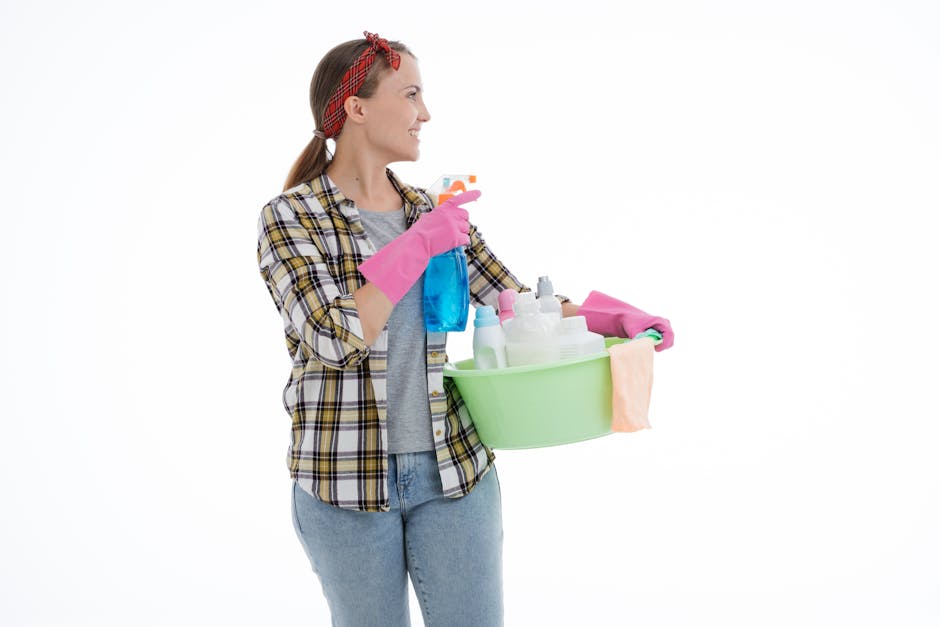 A woman with light skin and long brown hair, wearing a red headband, a plaid shirt, and jeans, is holding a green plastic basin filled with various cleaning supplies including spray bottles, cloths, and a sponge. She is also holding a blue spray bottle and a pink cloth, with a cheerful expression on her face, standing against a plain white background. This image represents surface cleaning and sanitisation, highlighting the tools and materials used in domestic cleaning, as part of the services offered by Hounslow Cleaner in their guide on high street domestic cleaning tips.