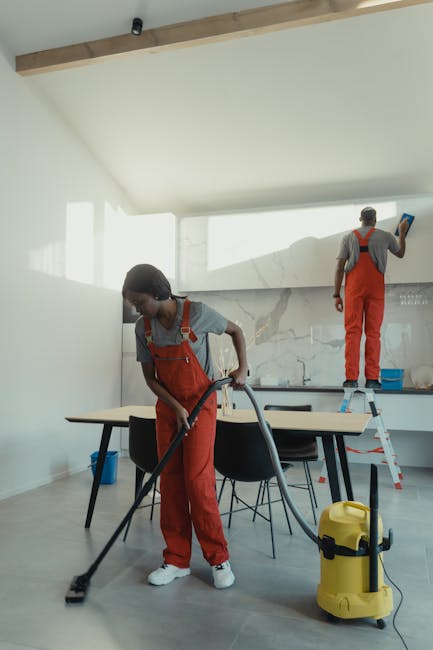 Two professional cleaners from Hounslow Cleaner are performing surface cleaning in a modern kitchen. One cleaner, a woman, is vacuuming the grey tiled floor with a handheld vacuum cleaner, while the o
