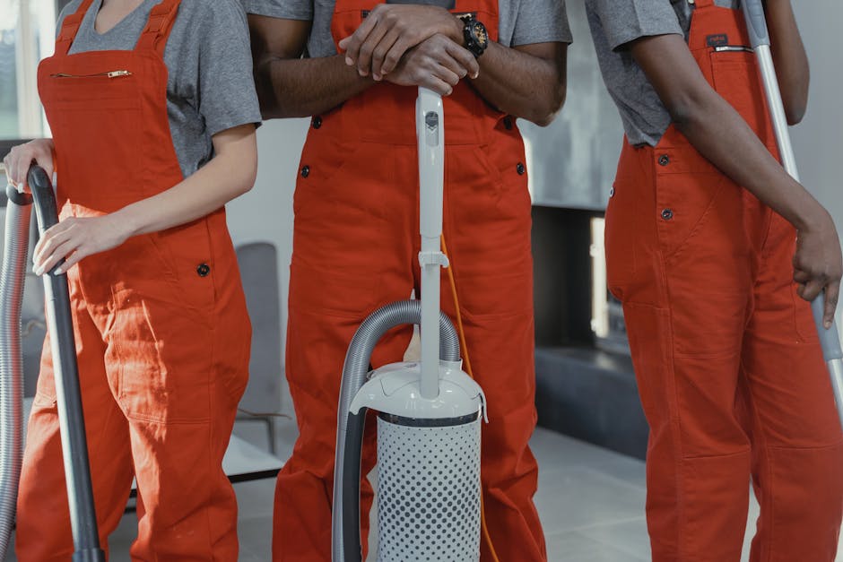 Two professional cleaners from Hounslow Cleaner are performing surface cleaning in a modern kitchen. One cleaner, a woman, is vacuuming the grey tiled floor with a handheld vacuum cleaner, while the o