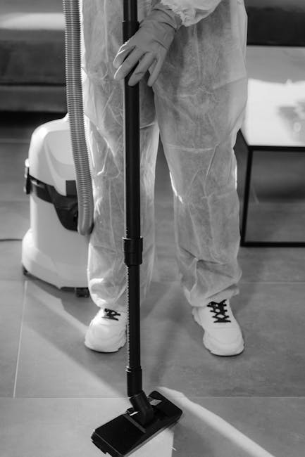 A person dressed in protective white coveralls and gloves is using a black handheld vacuum cleaner with a floor nozzle attachment to perform surface cleaning in an indoor setting. The vacuum cleaner is connected to a hose, which is attached to a larger cleaning machine placed behind the individual. The scene features a smooth, possibly wooden or laminate floor, with a portion of a dark-colored sofa or chair visible in the background. The bright lighting highlights the cleanliness of the floor, emphasizing the professional deep cleaning process. This image illustrates the routine maintenance and sanitisation practices employed by Hounslow Cleaner, supporting their offerings in domestic cleaning and end of tenancy cleaning around Hounslow.