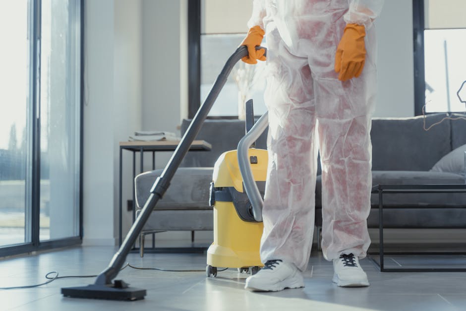 A professional cleaner dressed in protective white coveralls, orange gloves, and white sneakers is operating a yellow and gray commercial vacuum cleaner on a smooth, light gray tiled floor in a modern living room. The room features a large glass sliding door allowing natural light to illuminate the space, with a contemporary dark gray sofa and a wooden side table visible in the background. The surfaces, including the floor and furniture, appear spotless and well-maintained, reflecting thorough cleaning and sanitisation processes undertaken by Hounslow Cleaner as part of their end of tenancy cleaning services around Hounslow.